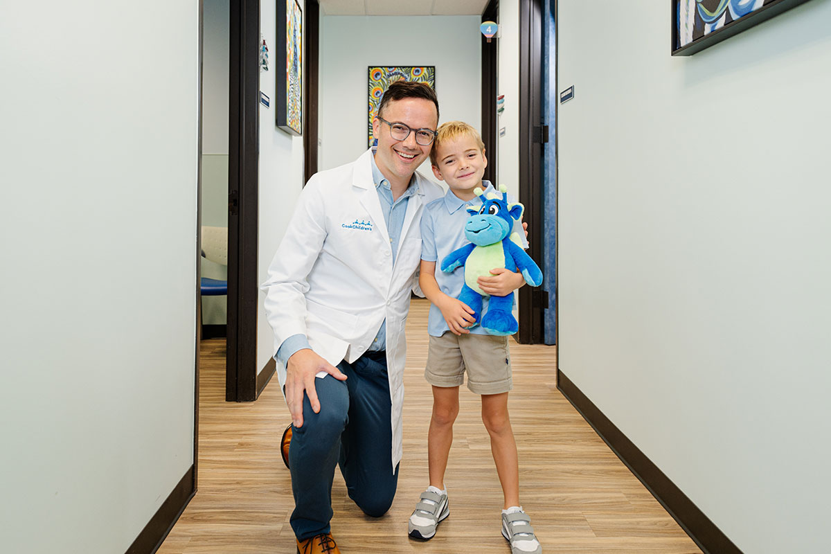 A pediatric physician and a child holding a stuffed dragon smile together at a pediatric primary care office