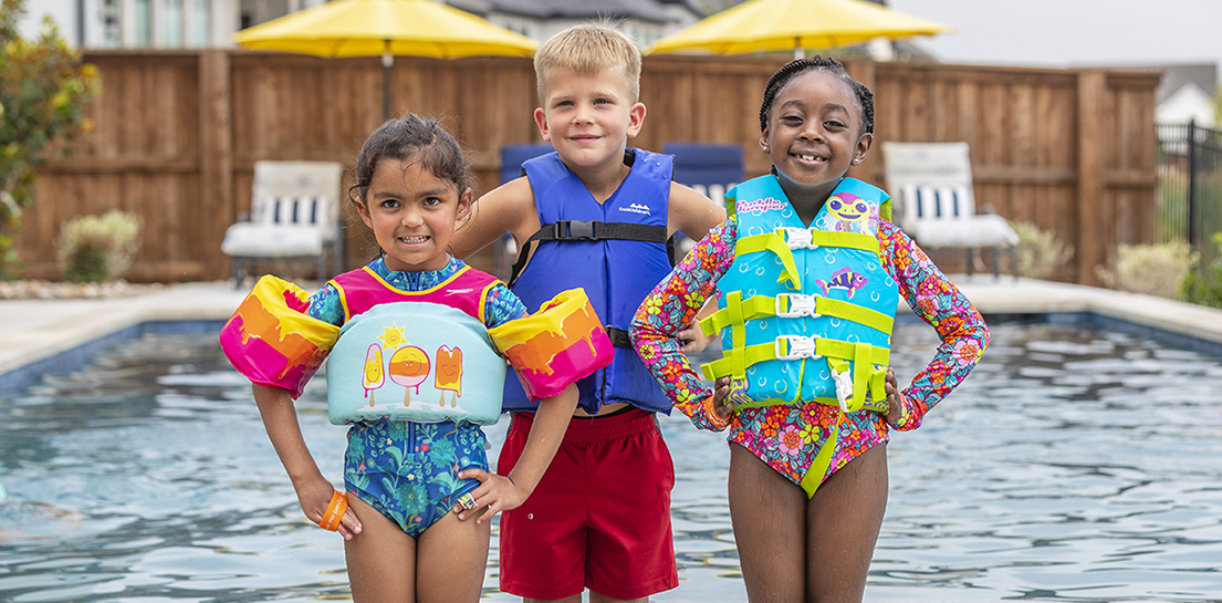 Three young children, standing next to backyard pool wearing coast guard approved, brightly colored lifejackets as recommended by Cook Children's water safety team