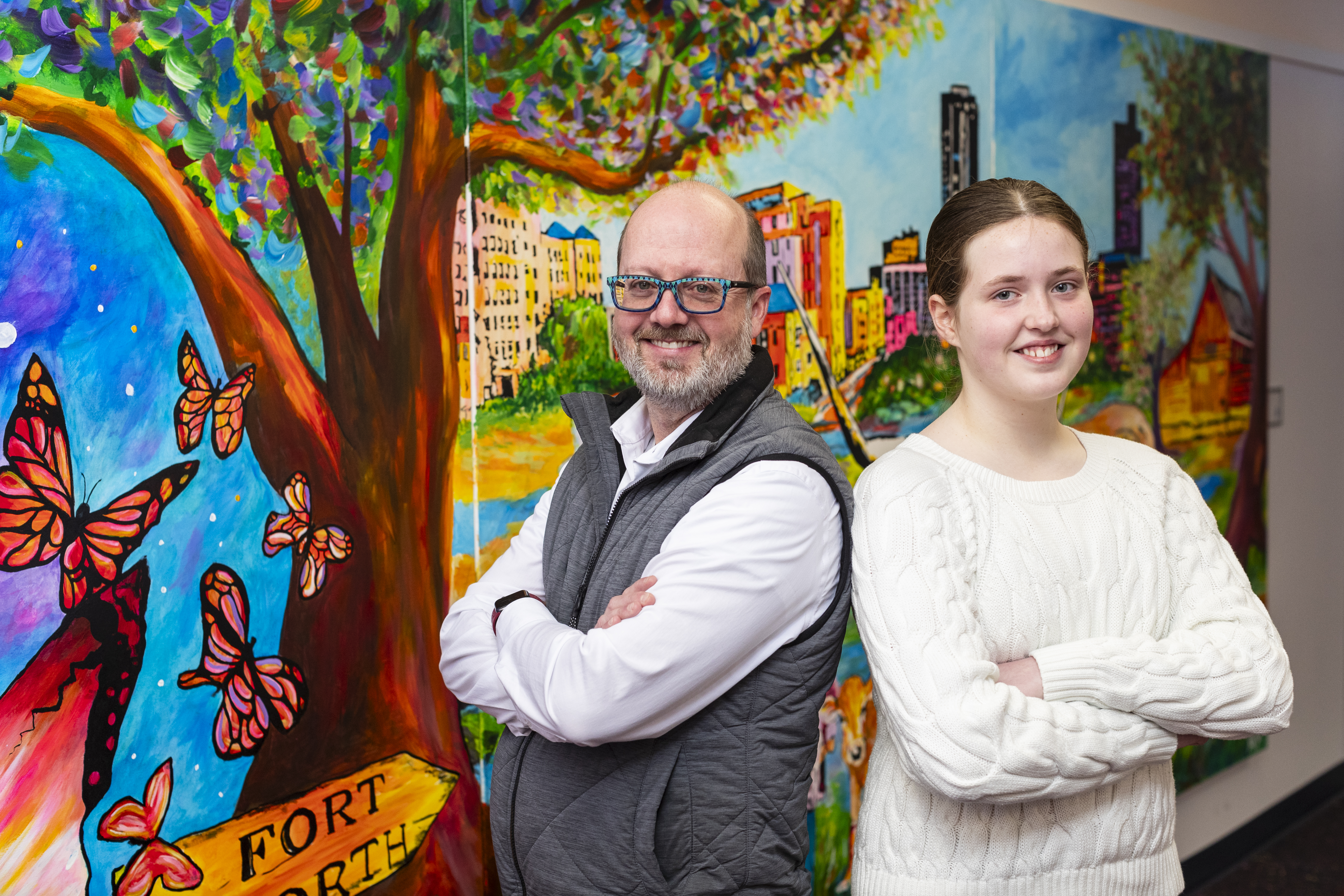 Doctor wearing blue glasses and a grey vest stands next to a young girl in a white sweater, they are in front of a colorful mural with butterflies