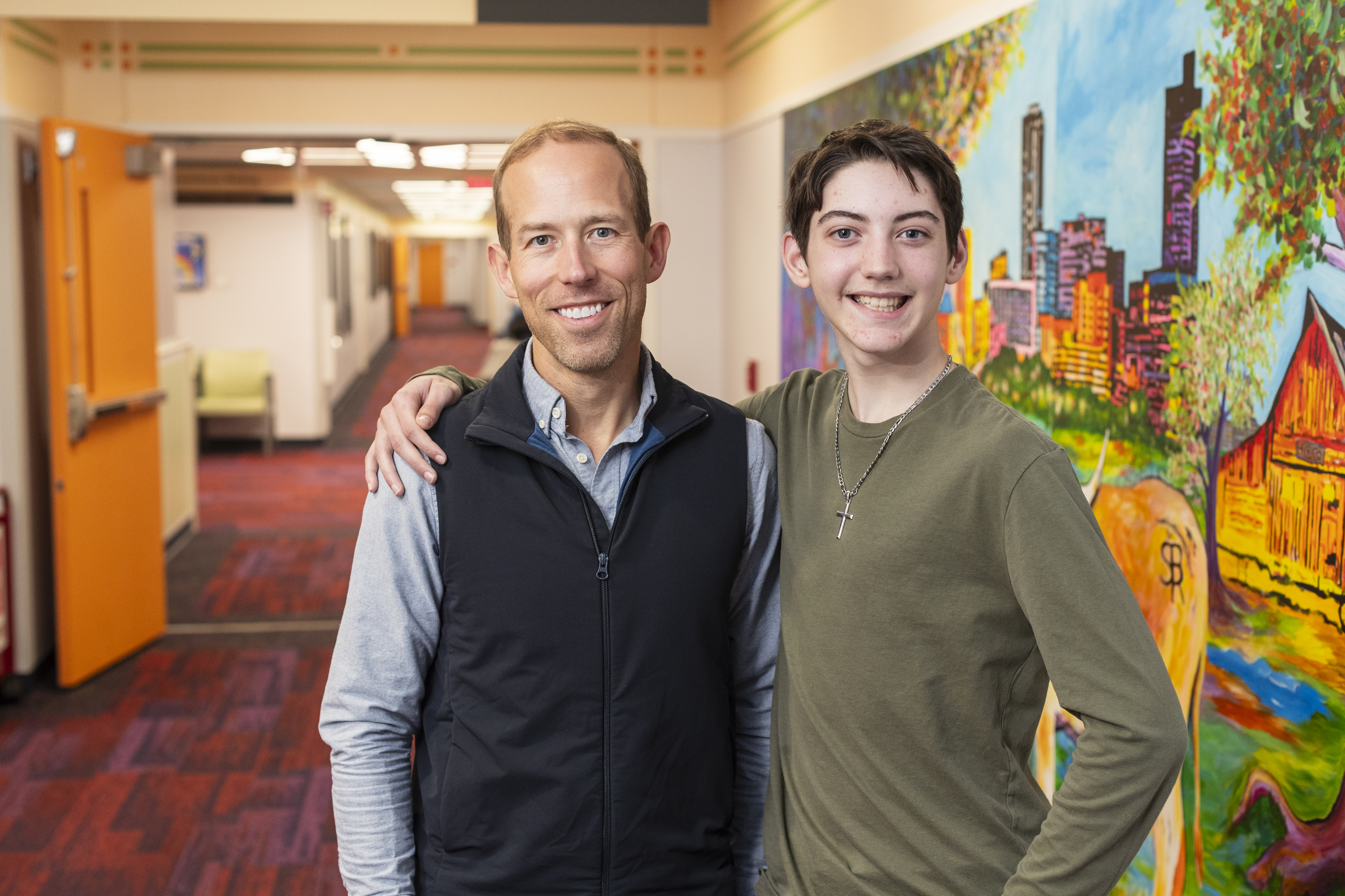 Young man wearing a green shirt has his arm around an older man wearing a blue shirt with a dark blue vest